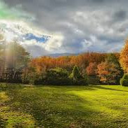 Campo gramíneo rodeado por árvores de outono, sob um céu nublado que contém raios de sol.